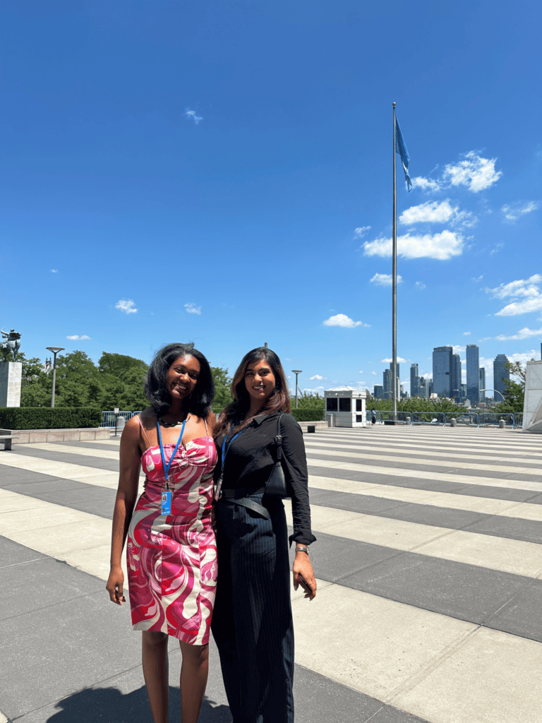 Anastasia Tomlinson and Samara Husain pose in front of the UN flag at the United Nations Headquarters in New York.